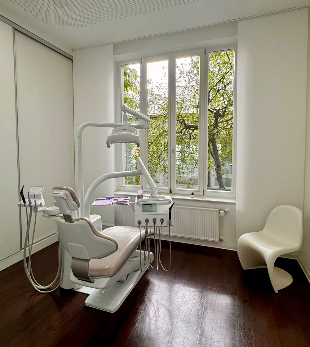 Heller Behandlungsraum mit weissem Zahnarztstuhl und Fenster mit Baumblick